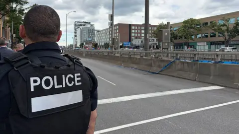 Back of officer in black vest marked with white letters reading "POLICE" next to an empty roadway with buildings across the street