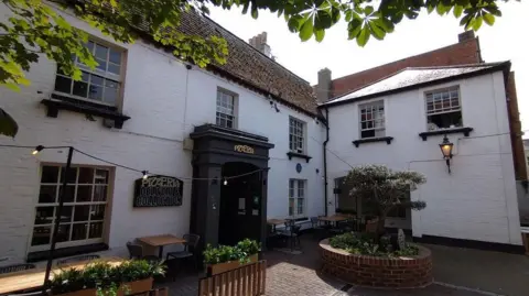 The entryway to the modern day Stanford Cottage. A sign on the black painted entrance reads 'PIZZERIA', now home to a Pizza Express. Tables and chairs, as well as potted shrubbery, line the open courtyard in front of the white painted building.