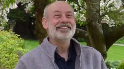 West Mercia Police A man with a beard stands outdoors wearing a jacket, with blossom trees in the background.
