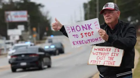 Reuters A man with his thumbs up on the side of the road tells motorists to "honk to stop executions" and "whoever is without sin cast the first stone". He wears a black jacket and ball cap, and cars are seen in the background