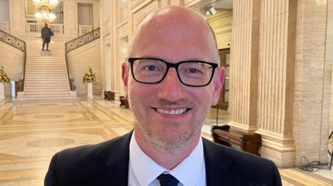 Peter Martin in a black suit white shirt and tie, smiling in Stormont's great hall, ornate walls and staircase in the background