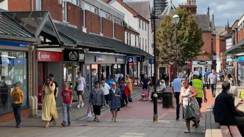 A busy town centre with people walking along a pedestrian street. There is a canopy on both sides above shop fronts.
