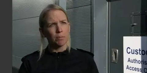 Ch Sup Baillie with long straight blonde hair wearing a black top and standing next to a door on which there is notice with "Custody Authorised Access" written in blue lettering.