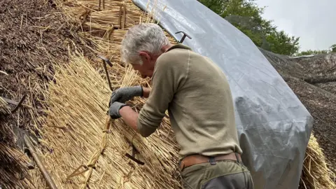 Alan Lewis is wokring on thatching a roof. He is up on a small step and working with the straw and metal tools. 