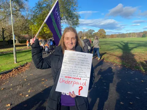 A woman waves a banner and holds a sign at a rally in a park.
