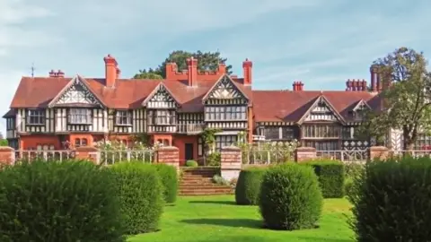 A large timber-framed building with tall brick chimneys and an ornamental garden with large bushes on a grass lawn