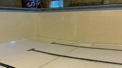 Ward Freman Community Pool Group An empty swimming pool, with cream tiles and black tiles, for the lane definition. There is a window above the pool, equipment in the corner and brick walls. 