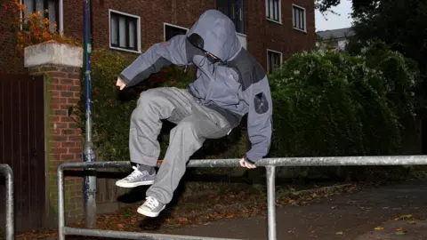 A generic picture of a teenage boy vaulting over a barrier by a house. His face is covered by the hood of his grey jacket and he has grey trousers and grey trainers.