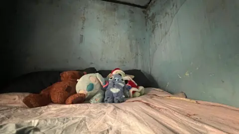 Emma Baugh/BBC News A bed with children's toys on it surrounded by a wall with water running down