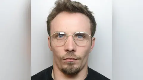 Thomas Harris mugshot. He has a goatee-like beard, moustache and is wearing glasses. He has quiffed brown hair.