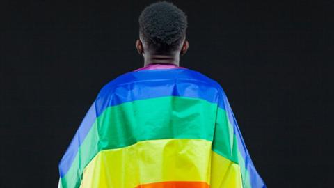 A man wrapped in a rainbow flag against a black background