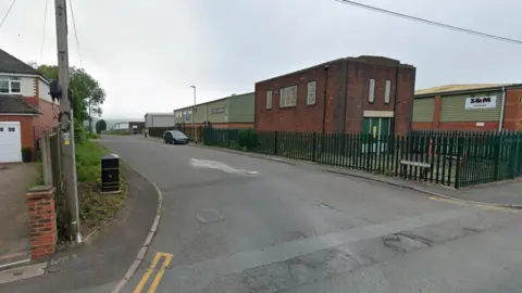 Google A general view of Canal Lane - a house is on the left and industrial buildings are behind a green fence on the other side of the road with a street sign that says "Canal Lane" in front of the fence.