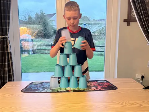 Logan McCarroll is a boy with blonde hair, he is standing in front of a set of glass patio doors wearing a blue top and is stacking light blue cups in a pyramid. He is standing in a living room with white walls.