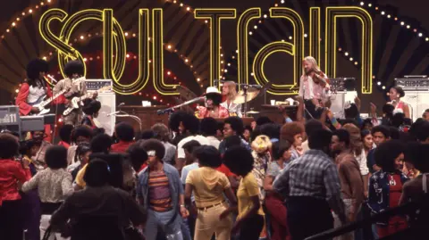 Getty Images A large sign that reads "Soul Train" in capitals hangs above a stage where Sly and the Family Stone perform to a dancing audience