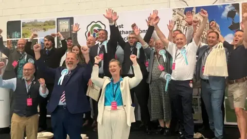 A group of mostly male councillors, some dressed in suits and ties and many having removed their jackets. They are holding their arms aloft celebrating their victory. There is a woman in a light-coloured jacket and trousers with a blue shirt occupying a prominent position at the front. Many are wearing light blue Reform UK rosettes. There is a West Northamptonshire Council banner behind them.
