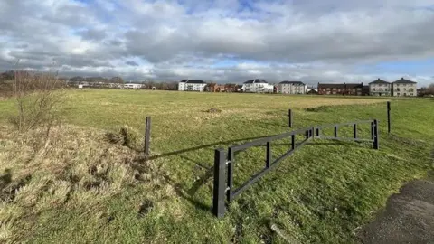 A large area of empty grass with housing in the background and part of a road to the right