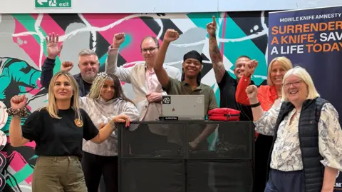 BBC Eight people stand next to a black podium with a laptop on top of it, with their hands raised in the air. Behind them is a multi-coloured background and a banner with a message about knife crime.