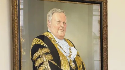 PA Media The portrait of former Belfast Lord Mayor Lord Wallace Browne on display at Belfast City Hall. The portrait is on a cream wall and in a gold frame.