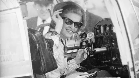A black-and-white image of a woman with a 1960s hairstyle wearing sunglasses and sitting in the cockpit of a small aircraft. She is smiling and waving at the camera.