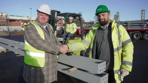 City of Wolverhampton Council Two men wearing a hard hat and hi-vis jacket are shaking hands and looking at the camera. They are near to two long steel objects.