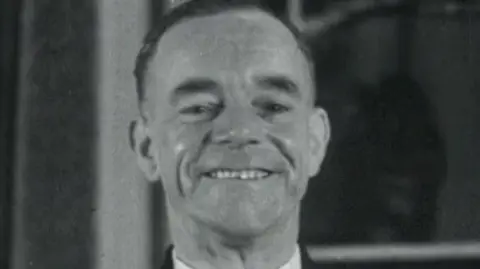 A screenshot from a black and white archive film from 1952.  Albert Gunter is smiling proudly and wearing his bus driver uniform.