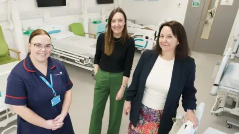 Left to right: SACT Clinical Educator at South Tyneside and Sunderland NHS Foundation Trust (STSFT) Rachel Boyd, STSFT Oncology Nurse Consultant  Lynsey Robson and programme leader Karen Humphreys smiling inside the University of Sunderland’s mock hospital ward. There are two hospital beds behind them with a green chair and a chest of drawers. 
