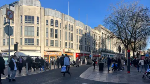 A busy pedestrianised shopping street with people everywhere and buildings behind, including a Pizza Hut and a Santander bank.