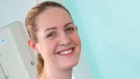 Lucy Letby smiles for the camera in a photo taken early in her career. She has long blonde hair in a ponytail and is standing in a hospital ward. 