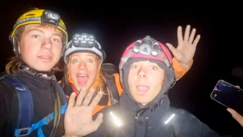 Family Photo Three individuals wearing helmets with headlamps and outdoor gear, posing for a photo in a dark environment. On the left of the picture is Draigen, who is 15, wearing a yellow caving helmet with a light on its front. He has light hair, which is long at the back. He has a dark blue fleece jacket, and has blue harness straps over his chest. To his left is mother Kate, staring into the camera with a wide open mouth and her left hand raised. She has a white caving helmet with a large light array on the helmet. She is wearing a bright orange coat and also has a harness or rucksack straps showing. To her left is Josh, 15. He is wearing a black coat with his hood up, and a pink caving helmet on the hood. He has both hands up waving, with a mobile phone in his left hand. The screen of the phone is a little cracked. Light from the phone taking the picture is lighting up reflective strips on shoulder of Josh's coat. It is pitch black behind them, with nothing visible, indicating they are underground.