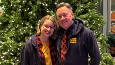 Lydia Greatrix Lydia Greatrix is looking at the camera and smiling. She is stood in front of a green Christmas tree and is wearing a yellow t-shirt, a red checked shirt and blue zip up hoodie. She has a blonde bob and wears glasses. Stood next to her is Kevin Tuerff who is looking at the camera and smiling and is wearing a blue Come From Away zip up hoodie and a red and black checked tie. 