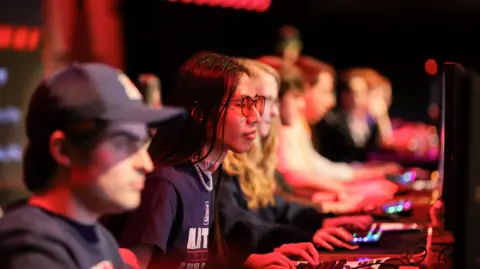 MEWC Players focused on their computers during a MEWC round in Las Vegas. The area is lit up with red light and only one person, along a line of people sat in front of computers, is in focus. They have long dark hair and are wearing tinted sunglasses.