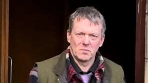 Mark Craster-Chambers is walking out of court, but is looking at the camera. He has short grey hair and a perplexed look on his face. He is wearing a green body-warmer, a red checked shirt and a purple tie.