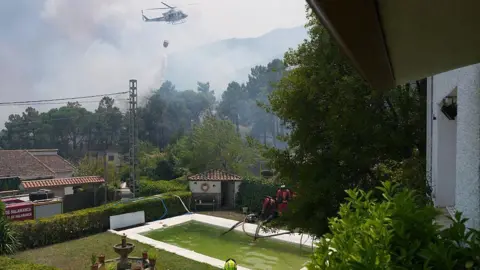 Cesar MANSO / AFP via Getty Images Firefighters pump water from a pool with a helicopter dumping water on a forest area filled with smoke