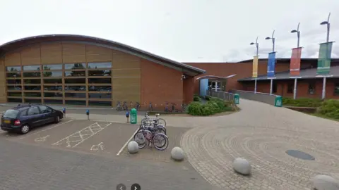 The front of White Horse Leisure and Tennis Centre in Abingdon. A large building with a curved roof is on the left, with the entrance to the right. Four large banners are on the bricked entrance ground in front of the car park.