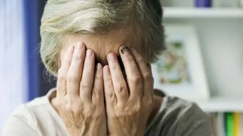 An elderly woman with white hair holds her face in her hands
