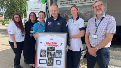 A man with short grey hair holds a rectangular display case containing a white shirt that says United Kingdom and has a UK flag at the top. Seven pictures are displayed underneath on the shirt. Four women wearing hospital uniforms stand next to him, along with a man wearing a short-sleeved shirt. 
