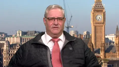 Doug Shipsey with grey hair is wearing glasses, a black coat, a tie and a white shirt. A London background, including Big Ben, is behind him.