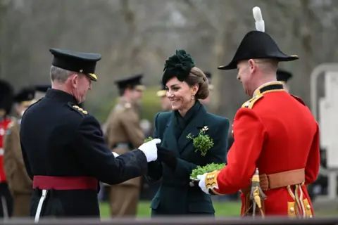 PA Media Princess of Wales wearing a green coat and hat with shamrock pinned, accompanied by a soldier in red uniform holding a tray of shamrocks while she presents a sprig of shamrock to a soldier wearing black.