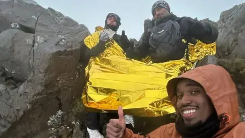 Nathan Mburu A selfie of three men sitting on the ridge of a foggy mountain. They all wear dark black, and grey clothing with the man taking the selfie in an orange coat. They all have their thumbs up. Two of the men have a foil blanket around them. 