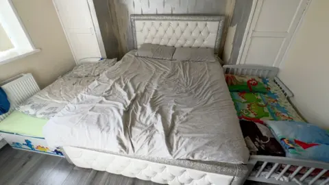 Camas em casa. Uma cama de casal fica entre um berço e uma cama de criança pequena. Há cobertores infantis na cama do berço e um desenho de dinossauro na cama das crianças. 