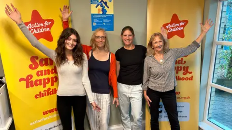 The four women stand infront of two yellow and red Action For Children banners, they are all smiling with their arms in the air and looking at the camera