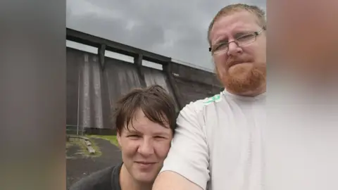 Social Media A selfie taken by Amanda and Christopher Fulton. On the left is Amanda, she has short brown hair and is wearing a grey t-shirt. She is resting her head on the arm of Christopher, who has short, light hair, a ginger beard and is wearing rectangle frame glasses and a white t-shirt. They are standing in front of a large, dark-coloured water reservoir. 