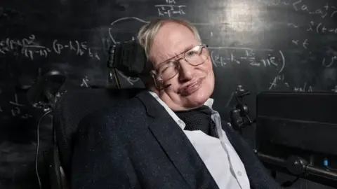 Richard Ansett/BBC Stephen Hawking, in a wheelchair, looking straight at the camera, wearing glasses, with a black board with white mathematical writing on it. He has a microphone by his mouth and a screen to his right. He is wearing a white skirt, with a cravat and a black jacket. 