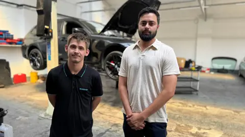 Ben Challice, on the left, with Amar Rehal. They are standing in a car workshop, with a silver can on a ramp behind them