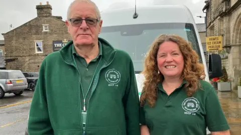 BBC/Seb Cheer A man and woman wearing green uniforms branded with "The Little White Bus" company logo stand in front of a minibus in Hawes.