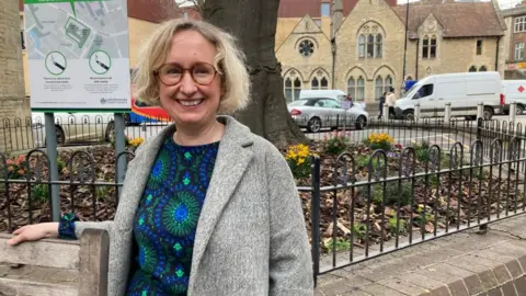 A woman stands in a colourful dress, wearing glasses. She looks happy.