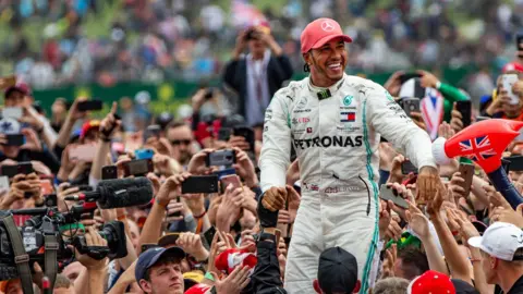 Craig Evans F1 driver Lewis Hamilton wearing a red cap and a white driving suit as he's held up high by a crowd