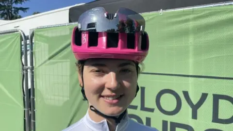 Alice Towers wearing a pink cycling helmet with sports sunglasses attached. She is wearing a white cycling jersey and stood in front of a green fence.