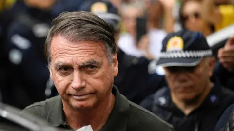 Reuters Jair Bolsonaro is wearing a dark green polo short as he leaves hospital on 14 September in Brasilia. A police officer can be seen behind him. 