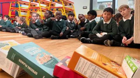 Up front, boxes of cereal having just fallen on top of one another. Behind, children sitting on the floor looking on.